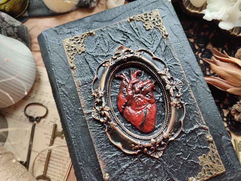 Pu&ograve; includere: Un diario rilegato in pelle nera con una cornice dorata ornata e un cuore anatomico rosso scolpito sulla copertina.