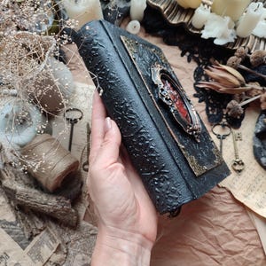 Pu&ograve; includere: Un diario rilegato in pelle nera con una chiusura in metallo decorata e un design rosso e dorato. Il diario &egrave; tenuto in una mano.