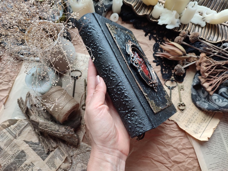 Pu&ograve; includere: Un diario rilegato in pelle nera con una chiusura in metallo decorata e un design rosso e dorato. Il diario &egrave; tenuto in una mano.