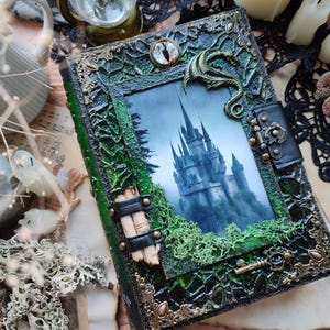 May include: A decorative, fantasy-themed journal with a green and black cover. The cover features a picture of a castle, a dragon, and ornate metal accents. The journal is embellished with moss and a key.