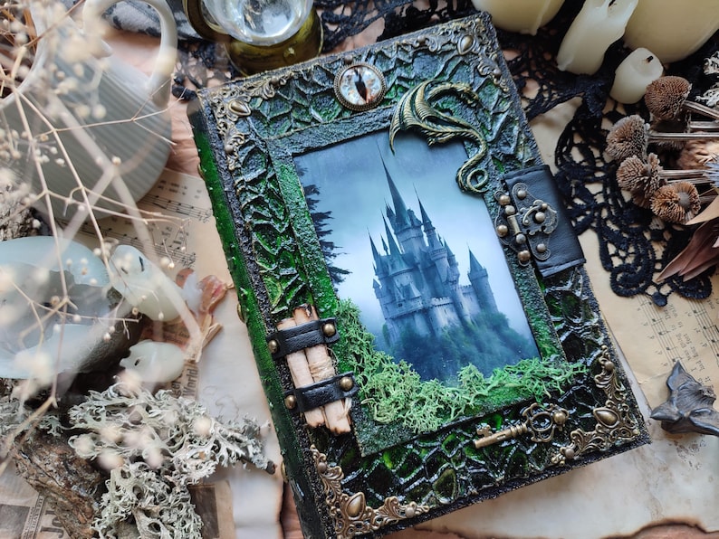 May include: A decorative, fantasy-themed journal with a green and black cover. The cover features a picture of a castle, a dragon, and ornate metal accents. The journal is embellished with moss and a key.