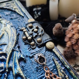 K&ouml;nnte beinhalten: Nahaufnahme eines blau-goldenen Fantasy-Journals mit Drachendesign. Das Journal hat einen dekorativen Verschluss, eine Kette, einen Lederriemen und Messingakzente. Der Einband ist aufwendig gestaltet.