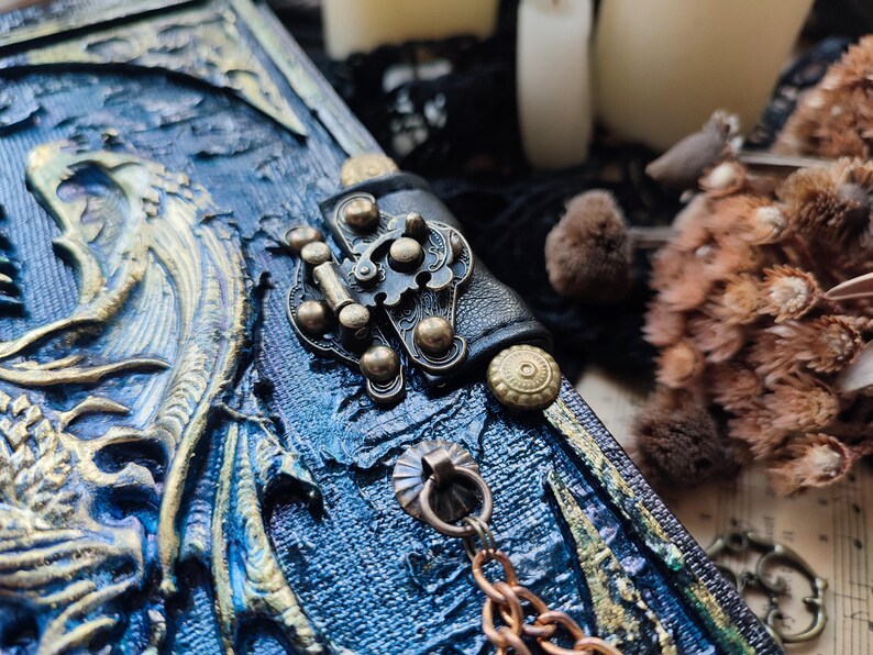 K&ouml;nnte beinhalten: Nahaufnahme eines blau-goldenen Fantasy-Journals mit Drachendesign. Das Journal hat einen dekorativen Verschluss, eine Kette, einen Lederriemen und Messingakzente. Der Einband ist aufwendig gestaltet.