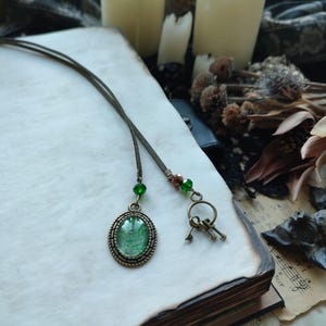 May include: A vintage-style necklace with a green oval pendant and small key charms. The pendant is set in a bronze-colored frame and hangs from a thin, dark cord. Green beads accent the cord. The necklace rests on an open book.