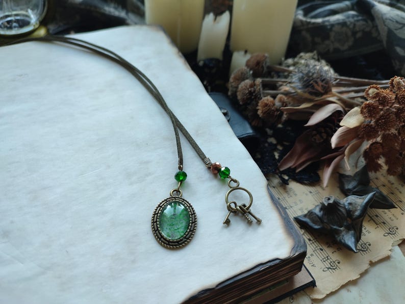 May include: A vintage-style necklace with a green oval pendant and small key charms. The pendant is set in a bronze-colored frame and hangs from a thin, dark cord. Green beads accent the cord. The necklace rests on an open book.