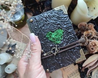 May include: A dark green and black journal with a textured cover and a green heart-shaped moss accent. The journal has brass corner protectors and a brown suede strap with a metal clasp. Decorative candles and dried flowers are in the background.