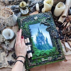 May include: A rectangular book with a dark green and black textured cover featuring a central image of a castle on a hill. The cover includes a small dragon ornament, a metal latch, a decorative key, and a single eye-shaped embellishment at the top.