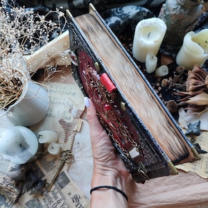 K&ouml;nnte beinhalten: Ein detailliertes, antikes Buch mit rotem und goldenem Einband, verziert mit aufwendigen Designs und einem Kettenverschluss. Die gealterten Seiten sind sichtbar, was auf ein Vintage- oder Fantasy-Thema hindeutet. Das Buch wird vor einem Hintergrund aus Kerzen und getrockneten Pflanzen gehalten.