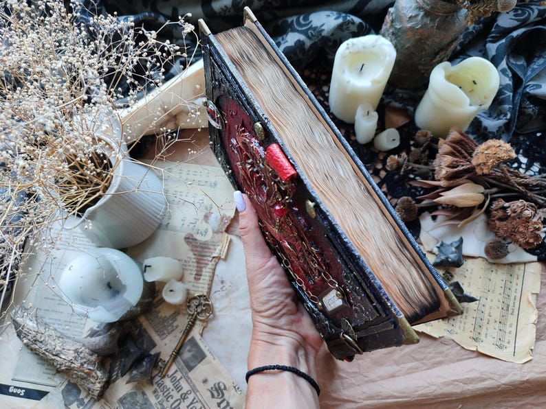 K&ouml;nnte beinhalten: Ein detailliertes, antikes Buch mit rotem und goldenem Einband, verziert mit aufwendigen Designs und einem Kettenverschluss. Die gealterten Seiten sind sichtbar, was auf ein Vintage- oder Fantasy-Thema hindeutet. Das Buch wird vor einem Hintergrund aus Kerzen und getrockneten Pflanzen gehalten.