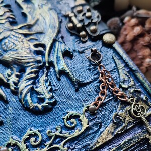 K&ouml;nnte beinhalten: Nahaufnahme eines blau-goldenen Fantasy-Journals. Der Einband zeigt ein Drachendesign mit komplizierten Details, eine Kupferkette und einen dekorativen Schl&uuml;ssel. Das Journal hat eine strukturierte Oberfl&auml;che.