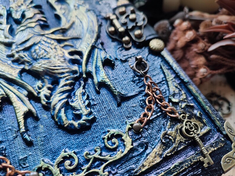 K&ouml;nnte beinhalten: Nahaufnahme eines blau-goldenen Fantasy-Journals. Der Einband zeigt ein Drachendesign mit komplizierten Details, eine Kupferkette und einen dekorativen Schl&uuml;ssel. Das Journal hat eine strukturierte Oberfl&auml;che.