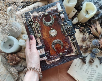 May include: A rectangular journal with a dark, textured cover featuring a central brown panel. The cover is decorated with two ornate metal crescent moon charms, a circular glass cabochon, three vintage-style skeleton keys, and a black latch.