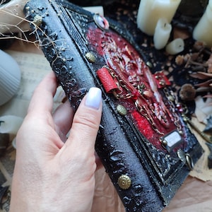 K&ouml;nnte beinhalten: Ein handgefertigtes Journal im gotischen Stil mit einem strukturierten schwarzen Einband und verzierten roten Akzenten. Das Journal verf&uuml;gt &uuml;ber Metallverzierungen, eine Kette und ein kleines Etikett. Der Buchr&uuml;cken hat gepr&auml;gte Details und messingfarbene Nieten.