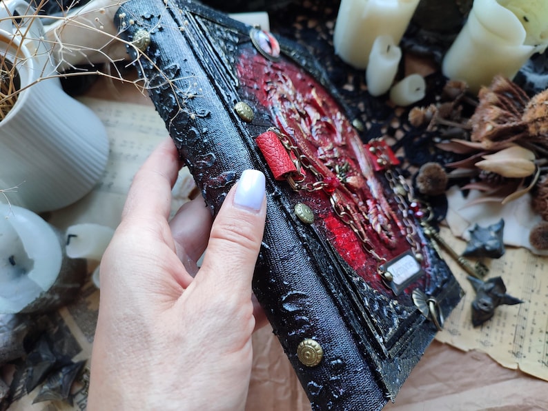 K&ouml;nnte beinhalten: Ein handgefertigtes Journal im gotischen Stil mit einem strukturierten schwarzen Einband und verzierten roten Akzenten. Das Journal verf&uuml;gt &uuml;ber Metallverzierungen, eine Kette und ein kleines Etikett. Der Buchr&uuml;cken hat gepr&auml;gte Details und messingfarbene Nieten.