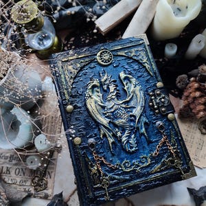 K&ouml;nnte beinhalten: Ein detailliertes, blau-goldenes Fantasy-Journal mit einem Drachenemblem. Das Cover hat ein strukturiertes, gepr&auml;gtes Design mit komplizierten Details, einschlie&szlig;lich eines Sonnen- und Mondsymbols. Dekorative Ketten und Verschl&uuml;sse tragen zur verzierten &Auml;sthetik bei. Das Journal ist von Kerzen und Schl&uuml;sseln umgeben.