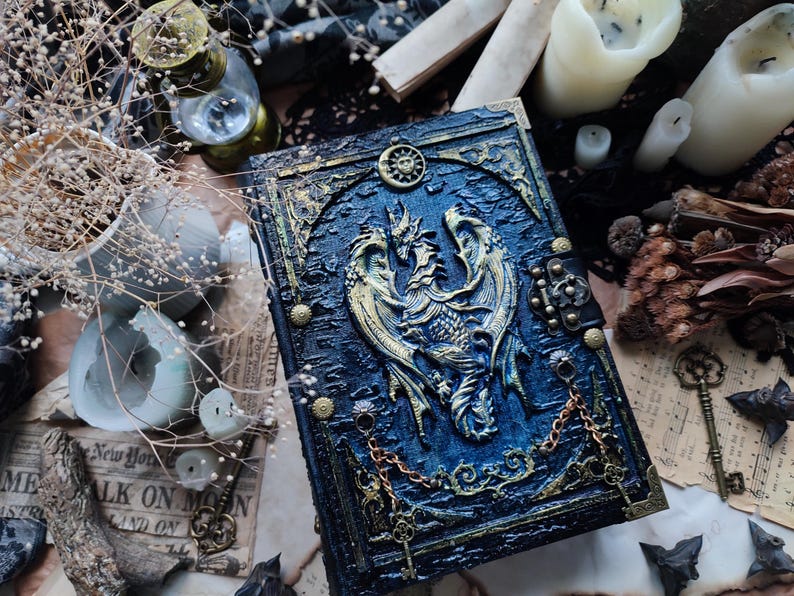 K&ouml;nnte beinhalten: Ein detailliertes, blau-goldenes Fantasy-Journal mit einem Drachenemblem. Das Cover hat ein strukturiertes, gepr&auml;gtes Design mit komplizierten Details, einschlie&szlig;lich eines Sonnen- und Mondsymbols. Dekorative Ketten und Verschl&uuml;sse tragen zur verzierten &Auml;sthetik bei. Das Journal ist von Kerzen und Schl&uuml;sseln umgeben.