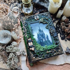 May include: A decorative, handcrafted journal with a fantasy theme. The cover features a detailed green and gold frame with a picture of a castle. A dragon and an eye adorn the cover. The journal is surrounded by candles and dried plants.