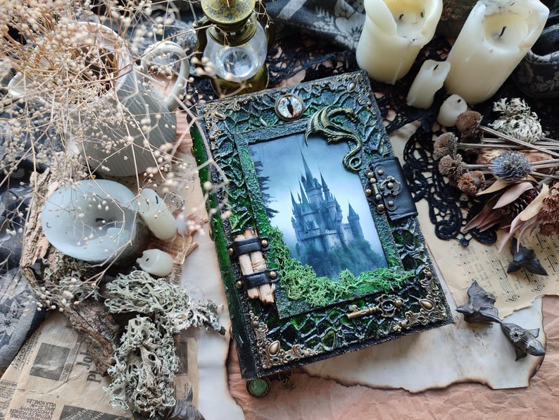 May include: A decorative, handcrafted journal with a fantasy theme. The cover features a detailed green and gold frame with a picture of a castle. A dragon and an eye adorn the cover. The journal is surrounded by candles and dried plants.