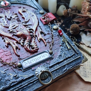 K&ouml;nnte beinhalten: Ein detailliertes, gotisches Tagebuch mit einem erhabenen Drachendesign in Rot und Gold. Das Tagebuch hat eine Metallplatte mit der Aufschrift "Memories", dekorative Ketten und einen Ringgriff. Die Gesamt&auml;sthetik ist dunkel und aufwendig.