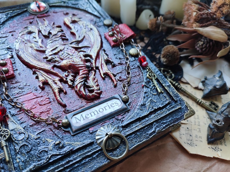 K&ouml;nnte beinhalten: Ein detailliertes, gotisches Tagebuch mit einem erhabenen Drachendesign in Rot und Gold. Das Tagebuch hat eine Metallplatte mit der Aufschrift "Memories", dekorative Ketten und einen Ringgriff. Die Gesamt&auml;sthetik ist dunkel und aufwendig.