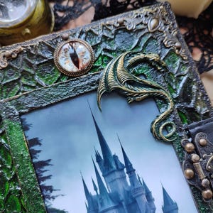 May include: A decorative journal with a green and bronze frame, featuring a castle image. The journal has a bronze dragon accent and an eye-shaped charm. The frame has a textured, ornate design and a black leather closure.