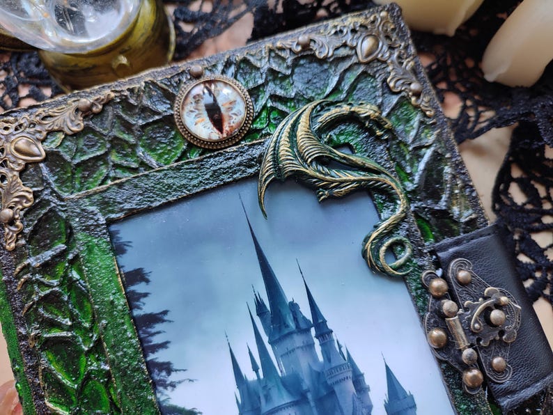 May include: A decorative journal with a green and bronze frame, featuring a castle image. The journal has a bronze dragon accent and an eye-shaped charm. The frame has a textured, ornate design and a black leather closure.