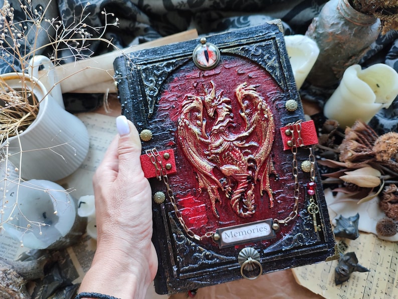 K&ouml;nnte beinhalten: Ein handgefertigtes Notizbuch mit einem roten und goldenen Drachendesign auf dem Einband. Das Notizbuch hat einen schwarzen Rahmen mit dekorativen Elementen, roten Lederriemen und einen Metallverschluss. Das Wort "Memories" ist auf einem kleinen Schild abgebildet.