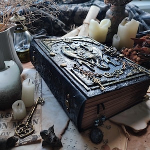 K&ouml;nnte beinhalten: Ein dunkelblaues, gepr&auml;gtes Buch mit kunstvollen goldenen Akzenten, Schl&uuml;sseln und einem Drachendesign. Das Buch ist von Kerzen, Trockenblumen und Vintage-Ephemera umgeben, was ein gotisches oder Fantasy-Thema erzeugt.