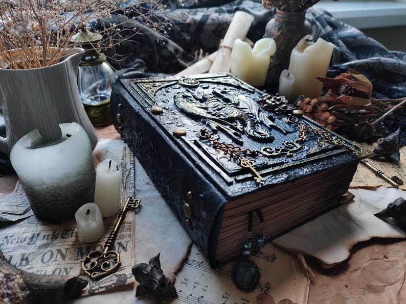 K&ouml;nnte beinhalten: Ein dunkelblaues, gepr&auml;gtes Buch mit kunstvollen goldenen Akzenten, Schl&uuml;sseln und einem Drachendesign. Das Buch ist von Kerzen, Trockenblumen und Vintage-Ephemera umgeben, was ein gotisches oder Fantasy-Thema erzeugt.