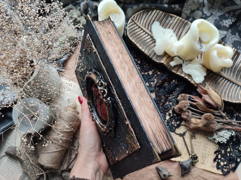 Pu&ograve; includere: Un diario rilegato in pelle nera con una cornice dorata decorativa e un design a forma di cuore rosso. Il diario &egrave; chiuso e ha un aspetto usurato e vintage.