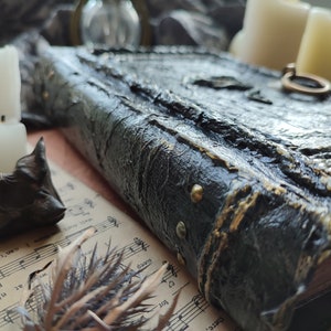 May include: A thick, rectangular book with a textured black cover featuring gold-colored highlights and metallic studs along the spine. A circular metal ring is attached to the front cover, which rests on a sheet of vintage sheet music.