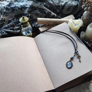 K&ouml;nnte beinhalten: Ein offenes, gealtertes Buch mit leeren Seiten, eine schwarze Kordelkette mit blauem Anh&auml;nger und Schl&uuml;sselanh&auml;nger sowie eine kleine Messinglaterne. Die Szene umfasst Kerzen, Trockenblumen und gerolltes Pergament.