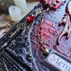 K&ouml;nnte beinhalten: Nahaufnahme eines dekorativen Journals mit rotem und schwarzem strukturiertem Einband. Ein bronzener Schl&uuml;sselanh&auml;nger und eine Kette mit roten Perlen h&auml;ngen an dem Journal. Das Wort "Mem" ist auf einem kleinen Metallschild sichtbar.