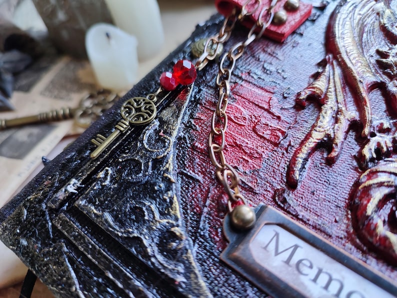 K&ouml;nnte beinhalten: Nahaufnahme eines dekorativen Journals mit rotem und schwarzem strukturiertem Einband. Ein bronzener Schl&uuml;sselanh&auml;nger und eine Kette mit roten Perlen h&auml;ngen an dem Journal. Das Wort "Mem" ist auf einem kleinen Metallschild sichtbar.