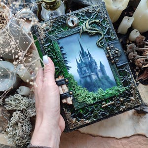 May include: A handcrafted, fantasy-themed journal with a castle illustration. The cover is dark green with ornate metal accents, a dragon embellishment, and a clasp. The journal's design evokes a mystical and gothic aesthetic.