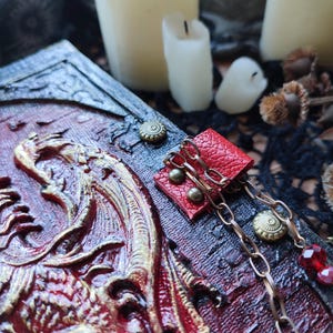 K&ouml;nnte beinhalten: Ein detaillierter, handgefertigter Buchumschlag mit einem Drachenrelief-Design in Rot und Gold. Der Umschlag hat ein abgenutztes, strukturiertes Aussehen. Ein kleiner roter Lederakzent mit Ketten- und Juwelen-Details ist angebracht.