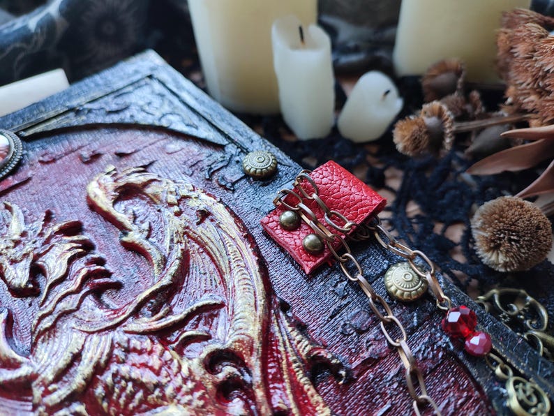 K&ouml;nnte beinhalten: Ein detaillierter, handgefertigter Buchumschlag mit einem Drachenrelief-Design in Rot und Gold. Der Umschlag hat ein abgenutztes, strukturiertes Aussehen. Ein kleiner roter Lederakzent mit Ketten- und Juwelen-Details ist angebracht.