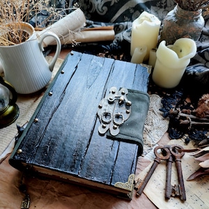 May include: A rectangular journal with a dark, textured wooden cover featuring vertical planks, metal corner protectors, and a central metal latch mechanism secured by a green fabric strap. The book is closed and rests on a flat surface.