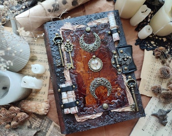 May include: A brown leather-bound journal with decorative metal accents, including keys, a lock, and crescent moons. The journal has a distressed, vintage look and is adorned with parchment paper and aged paper.