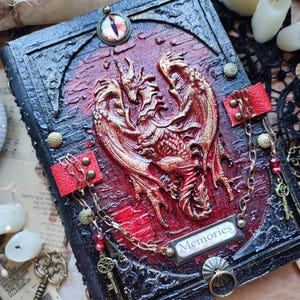 K&ouml;nnte beinhalten: Ein handgefertigtes Notizbuch mit einem roten und goldenen Drachendesign auf dem Einband. Das Notizbuch hat einen schwarzen Rahmen, rote Akzente und das Wort "Memories". Dekorative Ketten und Schl&uuml;ssel erg&auml;nzen das Fantasy-Thema.