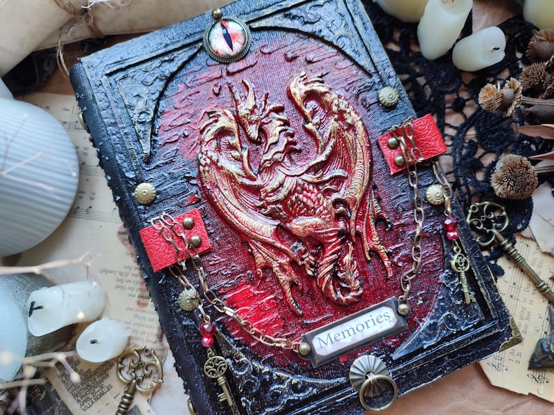 K&ouml;nnte beinhalten: Ein handgefertigtes Notizbuch mit einem roten und goldenen Drachendesign auf dem Einband. Das Notizbuch hat einen schwarzen Rahmen, rote Akzente und das Wort "Memories". Dekorative Ketten und Schl&uuml;ssel erg&auml;nzen das Fantasy-Thema.