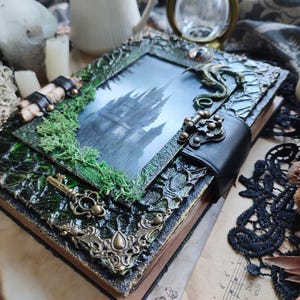 May include: A decorative, handcrafted journal with a gothic theme. The cover features a framed image of a castle, embellished with green moss, ornate metal accents, and a black leather strap. A vintage key is attached.