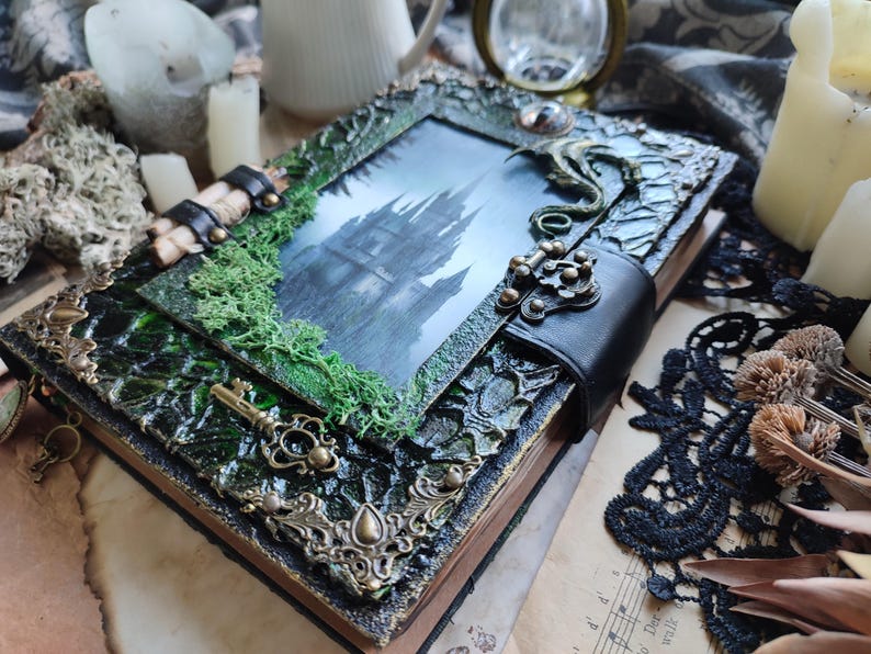 May include: A decorative, handcrafted journal with a gothic theme. The cover features a framed image of a castle, embellished with green moss, ornate metal accents, and a black leather strap. A vintage key is attached.