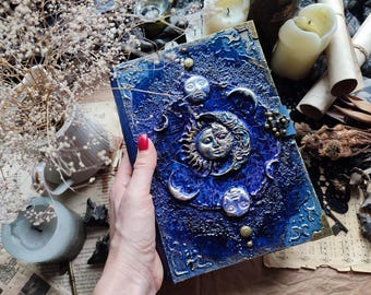 May include: A rectangular journal with a textured, deep blue cover featuring a prominent central sun and moon emblem. The cover is adorned with metallic silver and gold celestial motifs, including crescent moons and stars, with ornate brass-toned corner protectors.