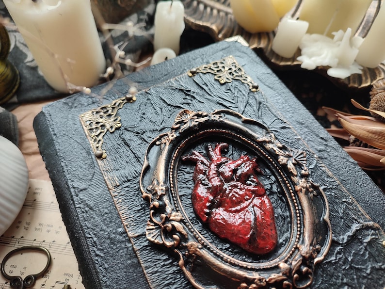 Pu&ograve; includere: Un diario rilegato in pelle nera con una cornice metallica decorativa che contiene un cuore anatomico rosso. Il diario ha un aspetto usurato e invecchiato ed &egrave; decorato con accenti dorati.