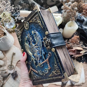 K&ouml;nnte beinhalten: Ein handgefertigtes Buch im Fantasy-Stil mit blauem Einband und Drachendesign. Das Buch hat goldfarbene Akzente, einen schwarzen Lederriemen und eine Kette. Die Seitenr&auml;nder sind sichtbar, und die Ecken sind mit goldenem Metall verst&auml;rkt.