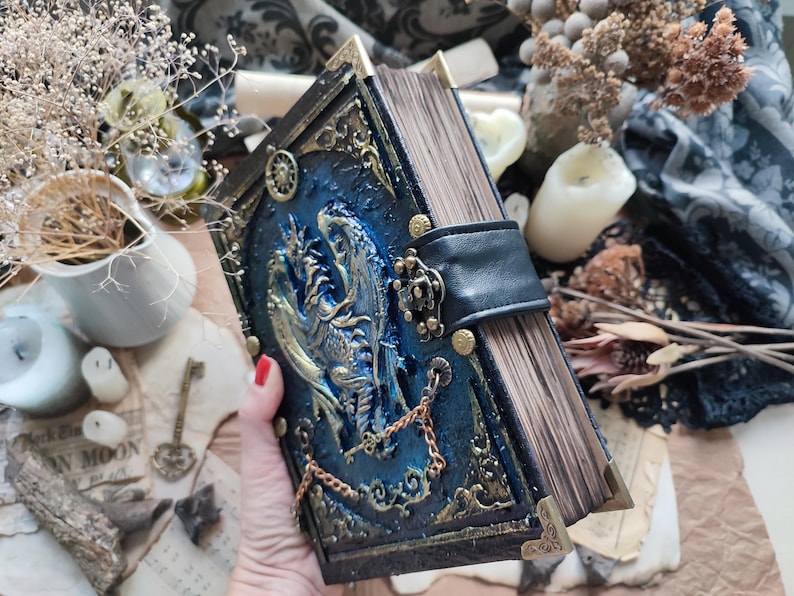 K&ouml;nnte beinhalten: Ein handgefertigtes Buch im Fantasy-Stil mit blauem Einband und Drachendesign. Das Buch hat goldfarbene Akzente, einen schwarzen Lederriemen und eine Kette. Die Seitenr&auml;nder sind sichtbar, und die Ecken sind mit goldenem Metall verst&auml;rkt.