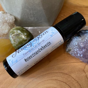 Może przedstawiać: Mała, przezroczysta butelka szklana z czarnym aplikatorem w postaci kulki. Butelka jest oznaczona etykietą "Nappy & Keen Essential Oil Blends #sneezinandwheezin DIRECTIONS".