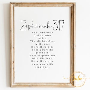 May include: A framed print featuring the text "Zephaniah 3:17" in a handwritten style. The text is a religious verse, printed in black on a white background. The frame is a light brown colour.