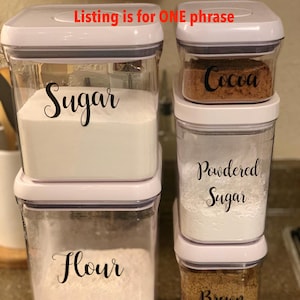 May include: Clear, rectangular food storage containers with white lids. The containers are labeled with black cursive text: "Sugar", "Flour", "Cocoa", "Powdered Sugar", and "Brown Sugar". The containers are filled with their respective ingredients.